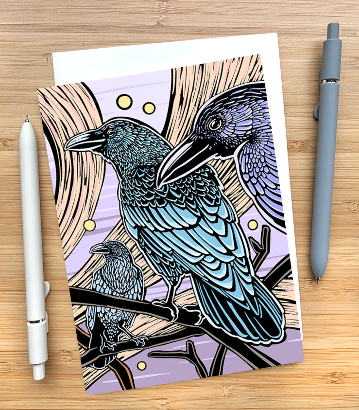 Card with a design of 3 crows against s purple sky with yellow swooshes on a wooden desk surface with two pens and an envelope.