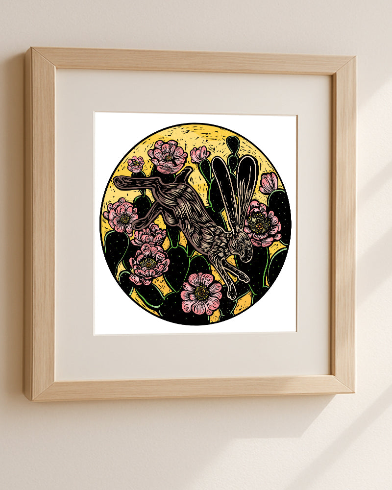 Framed artwork of a rabbit leaping over a prickly pear cactus on a white wall.