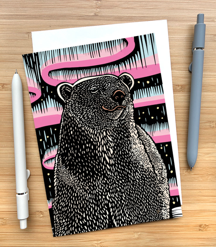 A greeting card featuring a woodcut style illustration of a polar bear set against a background with pink and light blue abstract aurora-like bands on a black field. The card sits on a wood surface with two pens beside it.