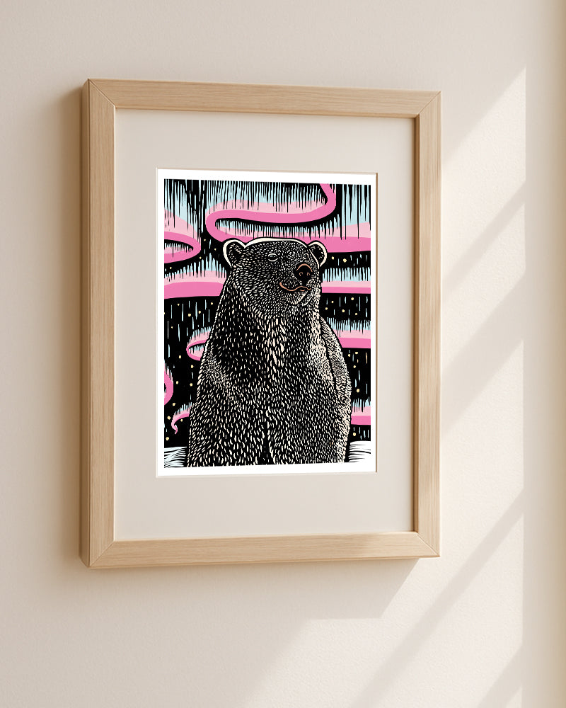 A framed art print featuring a stylized illustration of a polar bear, rendered in black and white linework with a magenta and light blue aurora background. The artwork is centered within a white border., displayed in a light wood frame on a beige wall.