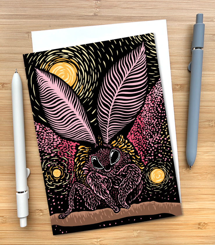 A greeting card featuring a woodblock style illustration of a rosy maple moth rendered in pinks and yellows, set against a black background with glowing yellow spheres. The card is styled on a wood surface with an envelope tucked inside and two pens.
