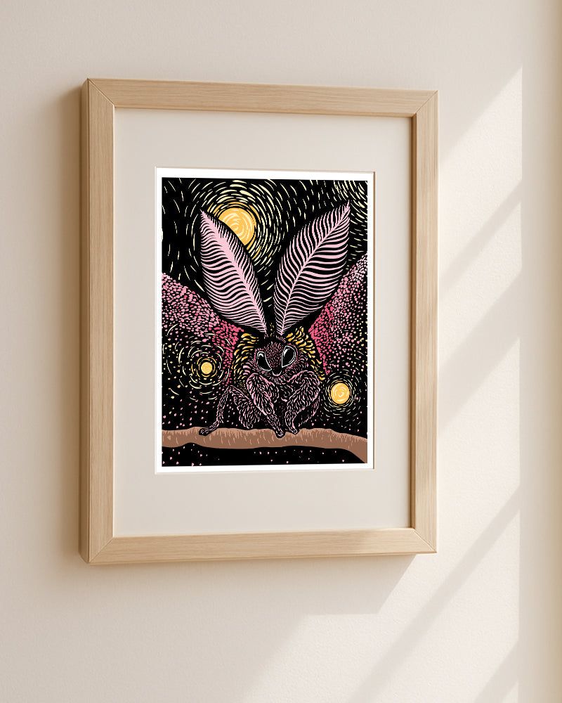A woodblock style print of a rosy maple moth, rendered in pink, black, yellow against a starry night sky with glowing spheres. displayed on a light beige wall in a natural wood frame with soft window shadows.