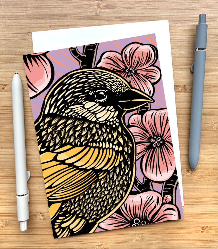 A greeting card featuring a woodcut-style illustration of a sparrow in front of cherry blossoms. The background is purple with pink streaks. The card sits on a wood surface and has two pens beside it.