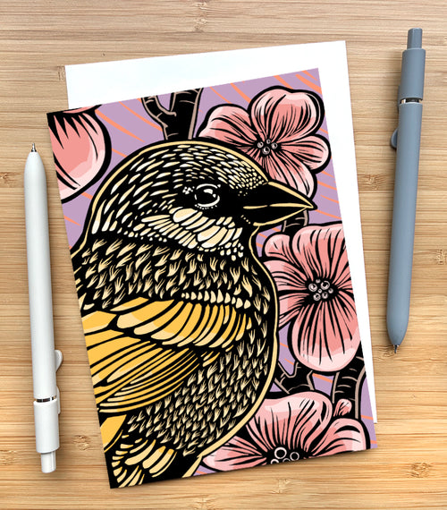 A greeting card featuring a woodcut-style illustration of a sparrow in front of cherry blossoms. The background is purple with pink streaks. The card sits on a wood surface and has two pens beside it.