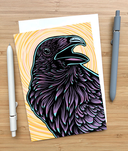 Notecard with a purple raven illustration on a wooden surface with two pens and an envelope.