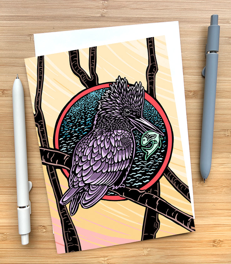 Card with a design of a kingfisher with a green fish in its mouth sitting on a branch with a stylized water and sky background on a wooden desk surface with two pens and an envelope.