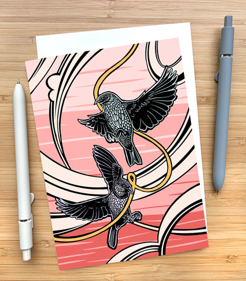 Card with a design of a two birds in flight with a pink sky background on a wooden desk surface with two pens and an envelope.