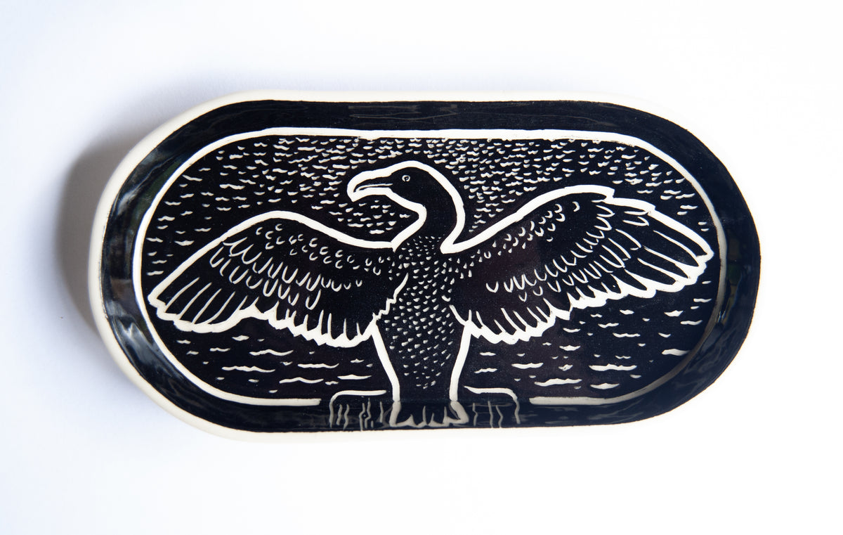 An oval black ceramic plate or tray featuring a carved image of a cormorant  with wings spread, set against a plain white background.