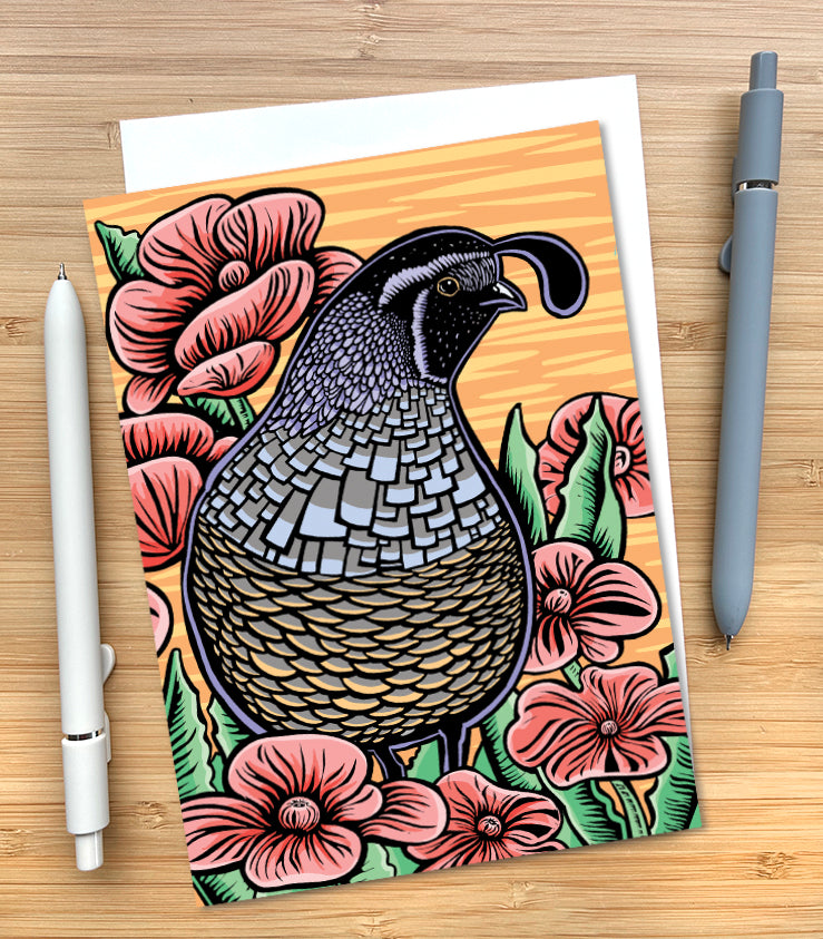 A greeting card featuring a woodblock style illustration of a California quail surrounded by pink flowers on an orange striped background. The card is styled on a wood surface with an envelope tucked inside and two pens. 
