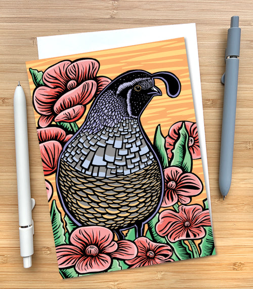 A greeting card featuring a woodblock style illustration of a California quail surrounded by pink flowers on an orange striped background. The card is styled on a wood surface with an envelope tucked inside and two pens. 