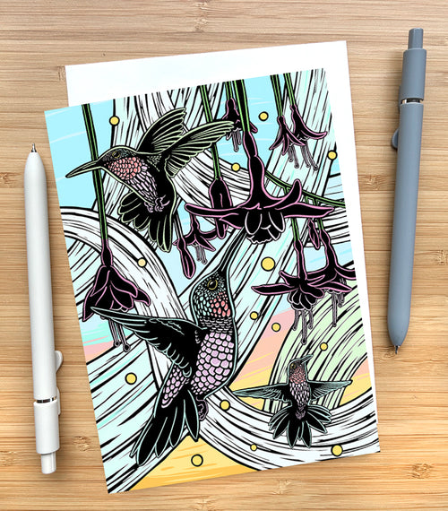 Card with hummingbird and flower illustration on a wooden surface with pens.