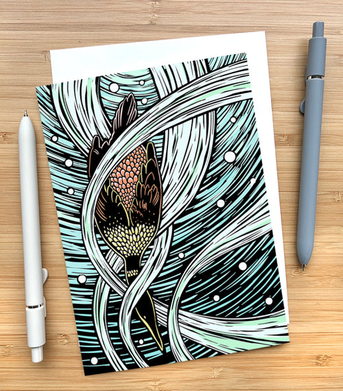Card with a design of a diving cormorant in a swirling water background on a wooden desk surface with two pens and an envelope.