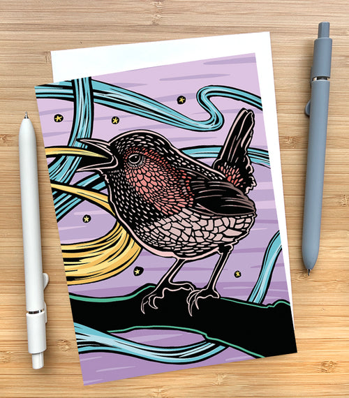 Card with a design of a wren sitting on a branch with a purple sky behind it on a wooden desk surface with two pens and an envelope.