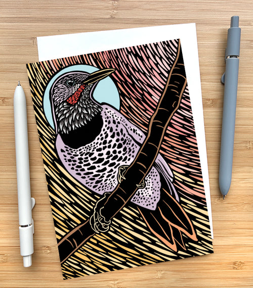 A greeting card featuring a stylized woodcut-style illustration of a flicker perched on a branch, with a light blue oval background behind the head and a pink and beige striped textured backdrop. The bird has black, white, and lavender-gray patterning with a small red accent near the beak. The card is sitting on a wood surface with two pens beside it.