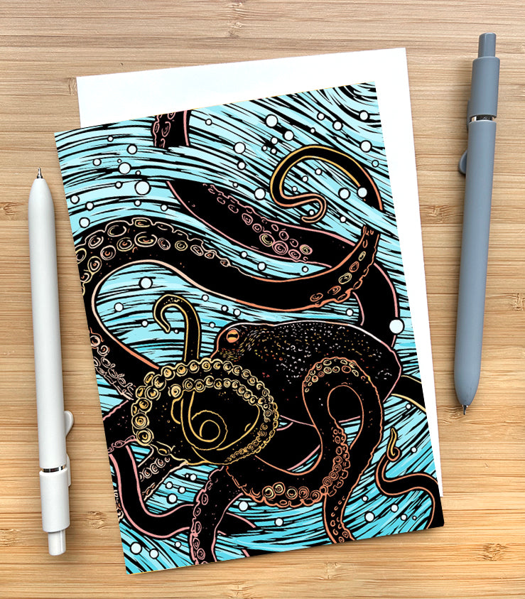 Notecard with octopus design on a wooden surface with two pens and an envelope.