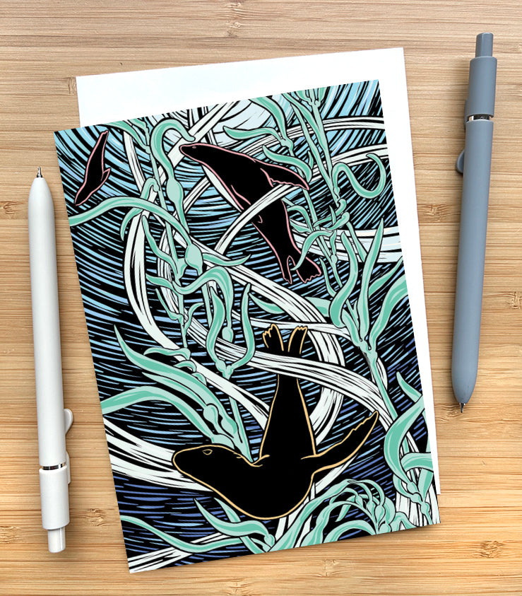 Card with a woodcut-style design of sea lions swimming through kelp a bird on a wooden desk surface with two pens and an envelope.