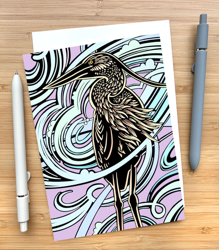 Card with a design of a heron with a swirling purple background sitting on a on a wooden desk surface with two pens and an envelope.