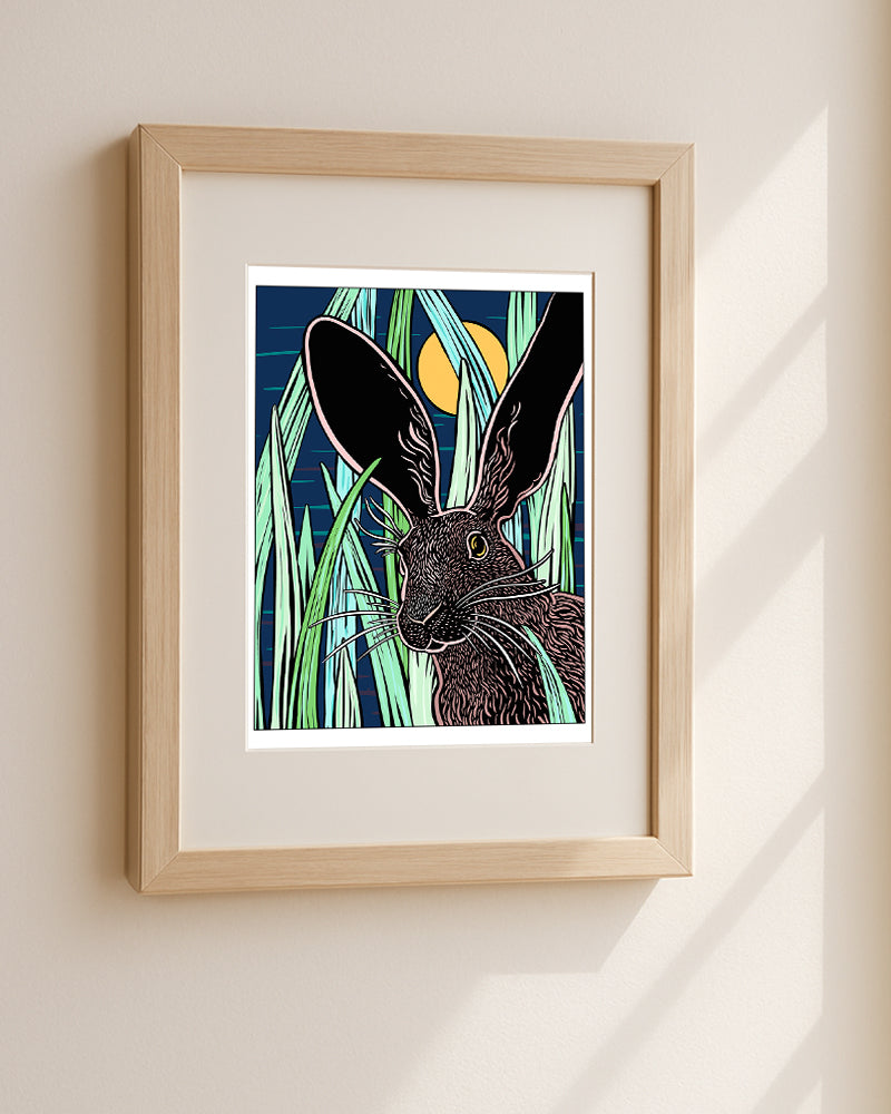Framed artwork of a rabbit in tall grass with a moonlit sky hanging on a white wall.