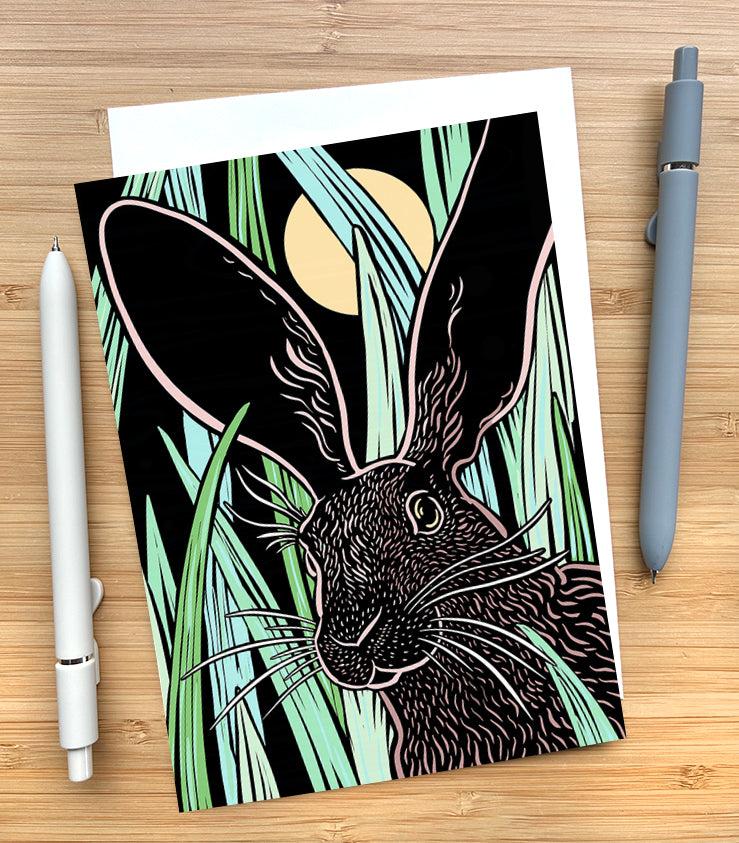 Greeting card with an illustration of a rabbit in front of green leaves sitting on a desk with two pens.