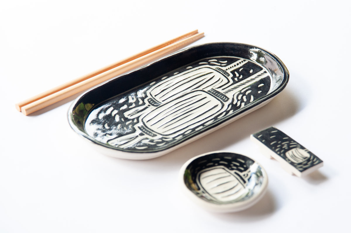 A set of black and white ceramic tableware featuring a hand-drawn lantern pattern, arranged on a white background alongside a pair of wooden chopsticks. The set includes a large oval platter, a small bowl, and a chopstick rest.