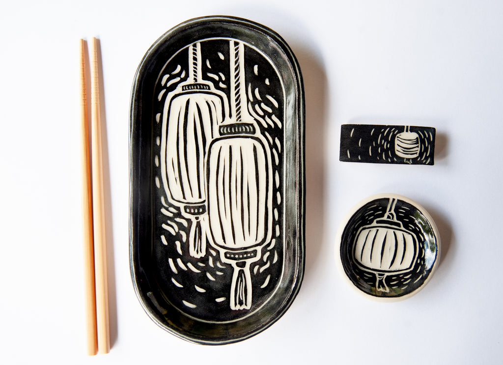 A set of black and white ceramic tableware featuring a hand-drawn lantern pattern, arranged on a white background alongside a pair of wooden chopsticks. The set includes a large oval platter, a small bowl, and a chopstick rest.
