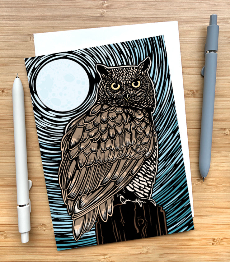 A greeting card featuring a detailed owl illustration with a large moon and swirling black linework on a blue-gray background, styled on a light wood surface with two pens beside it.
