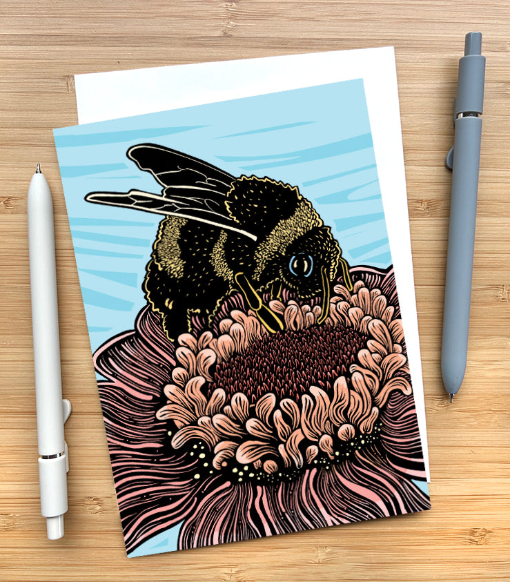 A 5x7 greeting card featuring a stylized woodcut style illustration of a bumblebee perched on a large pink flower. The card is sitting on a wood surface with 2 pens beside it.