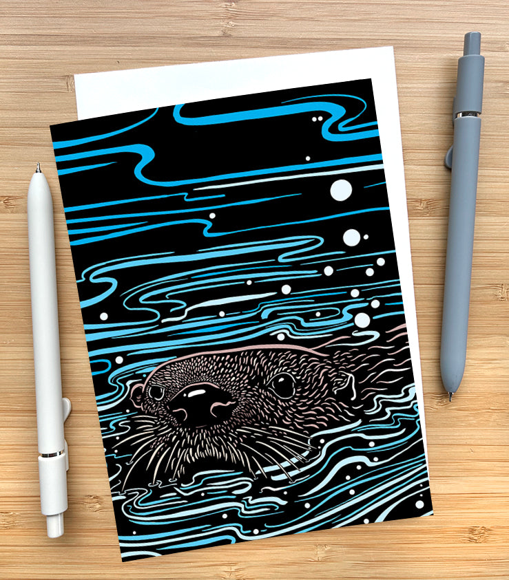 Card with a stylized otter illustration on a black background with blue and white patterns, placed on a wooden surface with two pens.