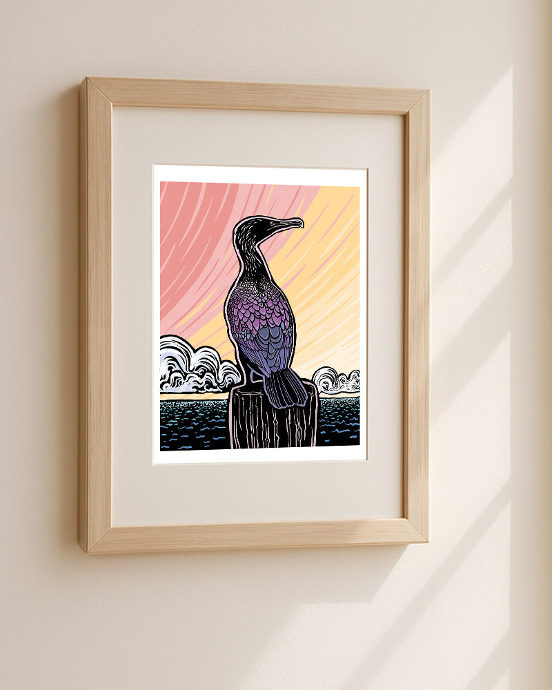 Framed artwork of a cormorant on a wooden pier with a colorful background hanging on a beige wall.