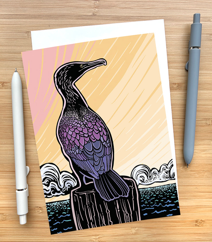 Card with a design of a cormorant sitting on a pier with a colorful sky behind them on a wooden desk surface with two pens and an envelope.