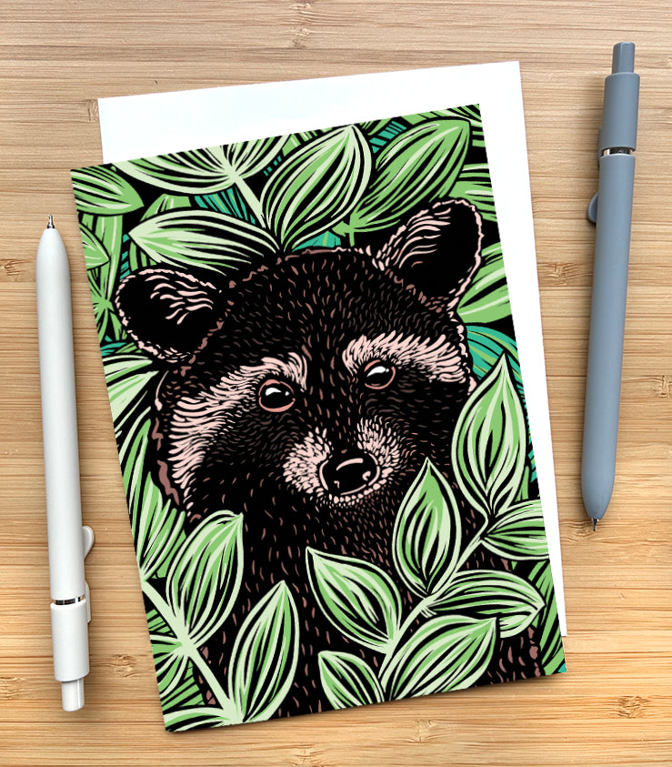 A card featuring a woodcut style illustration featuring a raccoon surrounded by green leaves. The card is on a wood surface with two pens beside it.