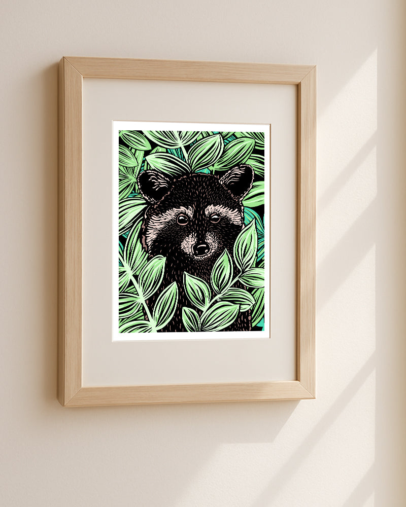A framed art print featuring a stylized illustration of a raccoon surrounded by bright green leaves, presented as an art print with a white border, displayed in a light wood frame on a beige wall with soft window shadows.