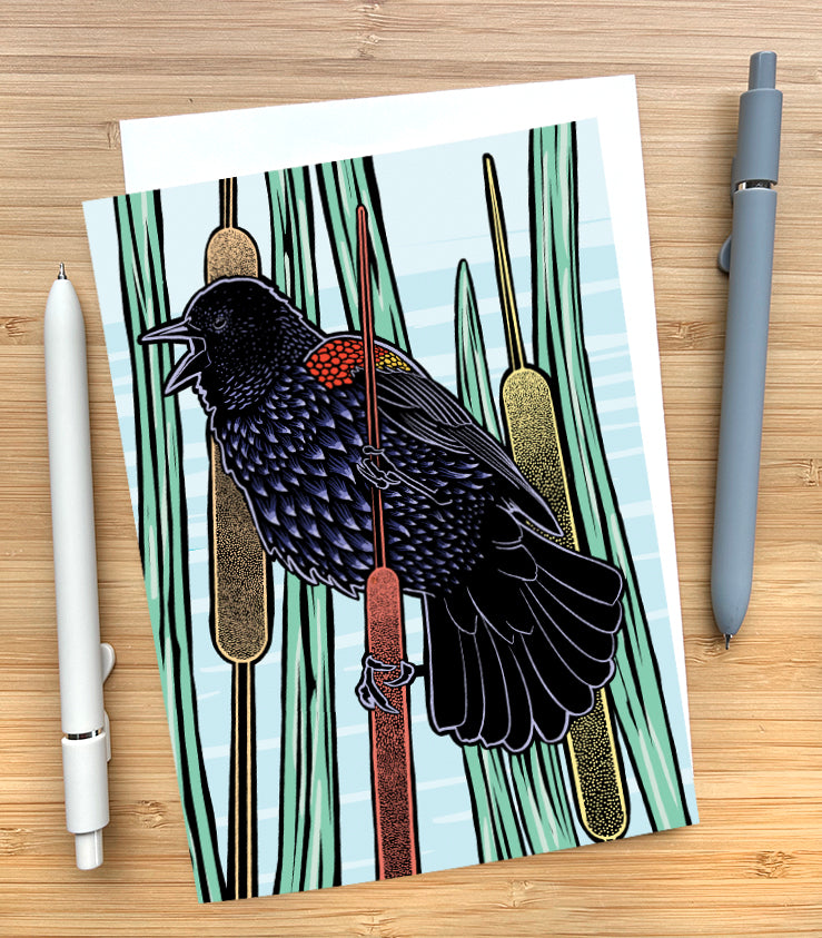 A card lying on a wood desk featuring a block-print style image of a red-winged black bird on a cattail plant. More cattails are in the background and the sky behind them is blue. 