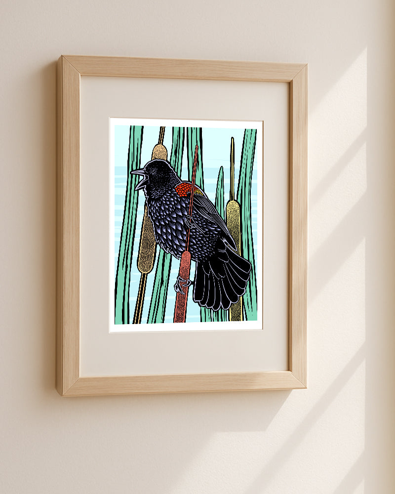 Framed art print featuring a red wing black bird with perched among tall green reeds, displayed in a light wood frame on a beige wall with soft sunlight shadows.