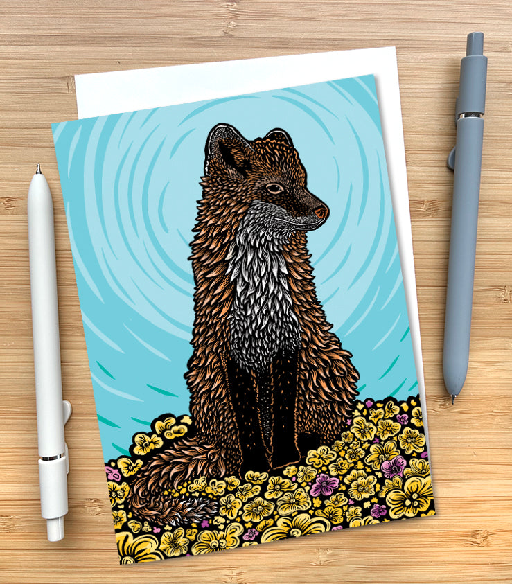 A greeting card featuring a thoughtful red fox sitting in a field of yellow and magenta flowers against a blue background.The card is displayed on a light wood surface, styled with a white pen on the left and a gray pen on the right.