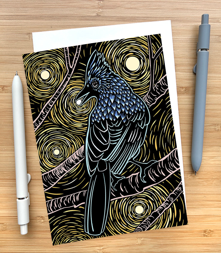 Card with a design of a stellar’s jay sitting on a branch with the moon behind them on a wooden desk surface with two pens and an envelope.