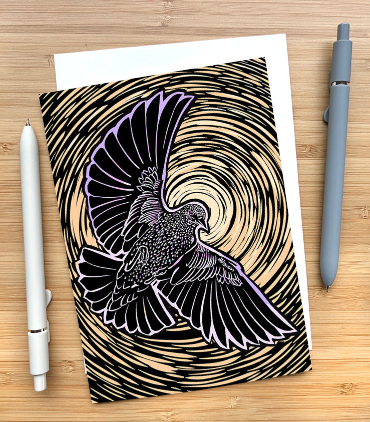 Card with a design of a starling flying with a yellow circular design behind them on a wooden desk surface with two pens and an envelope.