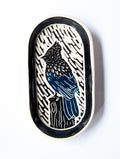 An oval ceramic tile featuring a black and blue linocut-style illustration of a bird perched on a tree stump, set against a white background.