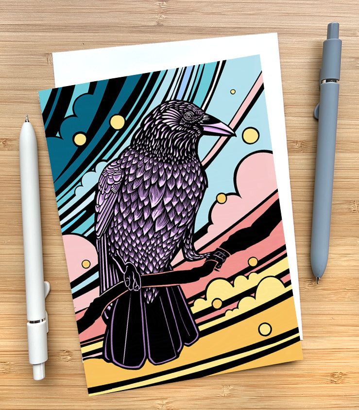 Card with a design of a crow sitting on a branch with a colorful sky on a wooden desk surface with two pens and an envelope.