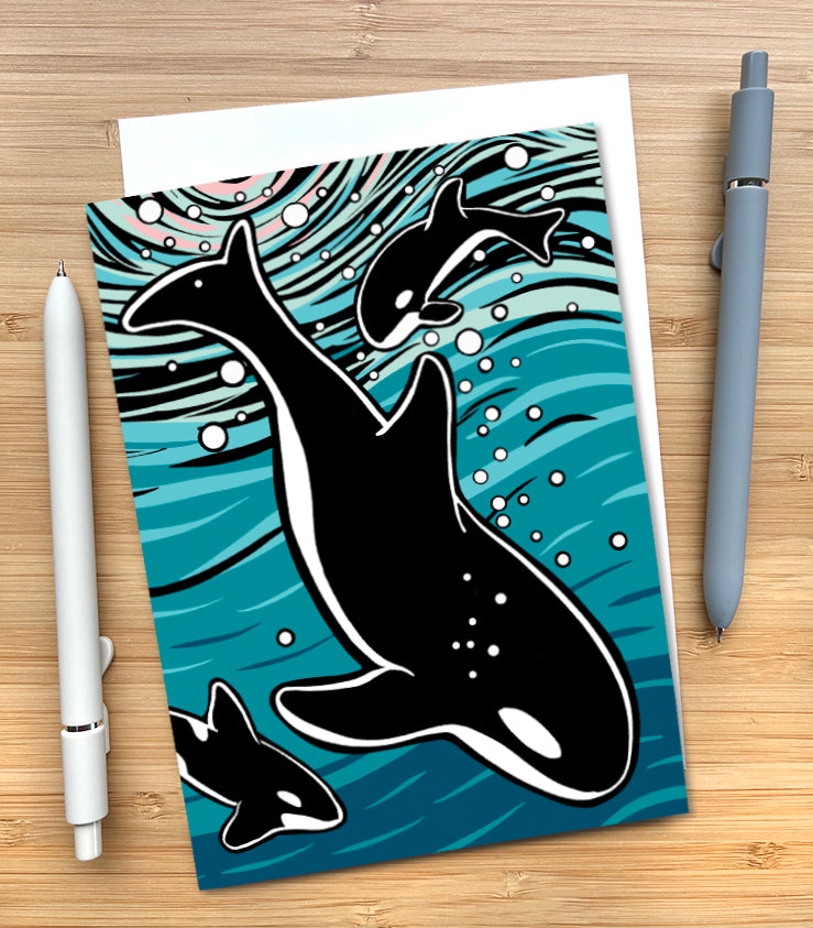 A greeting card featuring a stylized illustration of three orcas swimming in teal and blue ocean waves, styled on a light wood surface with two pens beside it.