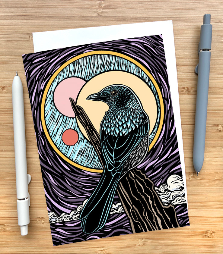 Card with a design of a bird sitting on a branch with the moon behind them on a wooden desk surface with two pens and an envelope.